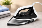 A modern steam iron with a white and black body, resting on a light marble surface with folded blue and gray clothes and a small potted plant in the background.