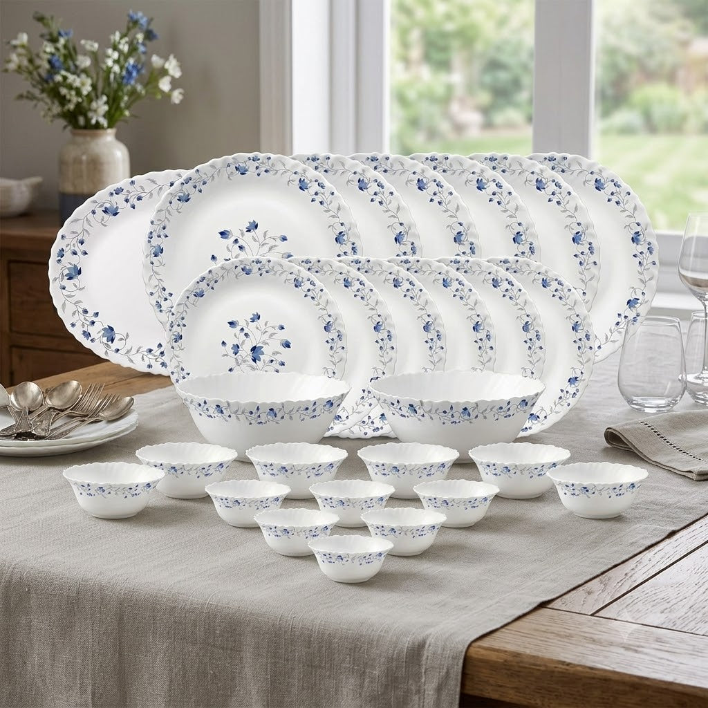 A coordinated dinnerware set arranged on a table in a real dining room setting. The set includes multiple white plates, bowls, and cups with a delicate blue floral pattern around the rims and centers.