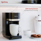 A black Lifelong coffee maker with a silver accent band sits on a white marble countertop with a white mug placed underneath. A second mug, a plate of cookies, and a white canister are visible in the background.