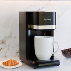 A black coffee maker with a silver band and Arabic script logo, dispensing coffee into a white ceramic mug on a marble countertop, with a croissant and coffee beans nearby.