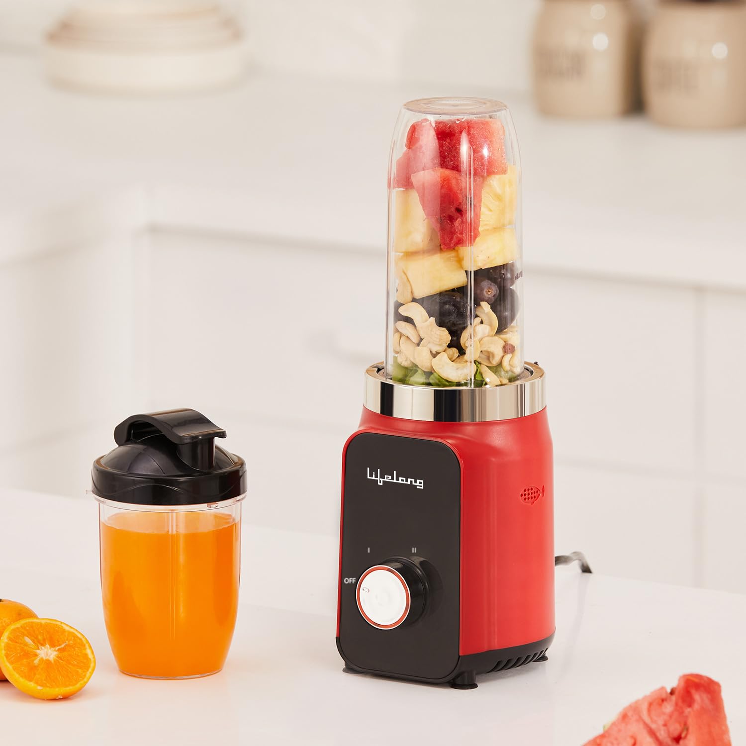 Portable personal blender with a red motor base and smoothie cup, blending fresh watermelon, pineapple, blueberries, cashews, and greens in a modern kitchen. Includes a travel cup filled with fresh orange juice, ideal for smoothies, protein shakes, and healthy drinks.