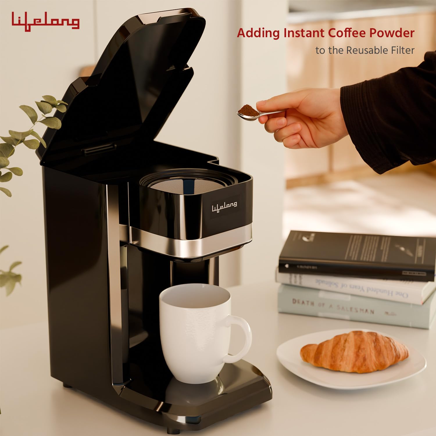 A black Lifelong single-serve coffee machine with its lid open, shown on a countertop with a white mug underneath. A hand is adding instant coffee powder from a small spoon into the reusable filter basket.