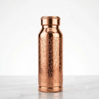 A metallic copper-colored bottle with an ornate engraved floral pattern, standing upright on a light marble surface against a white studio background.