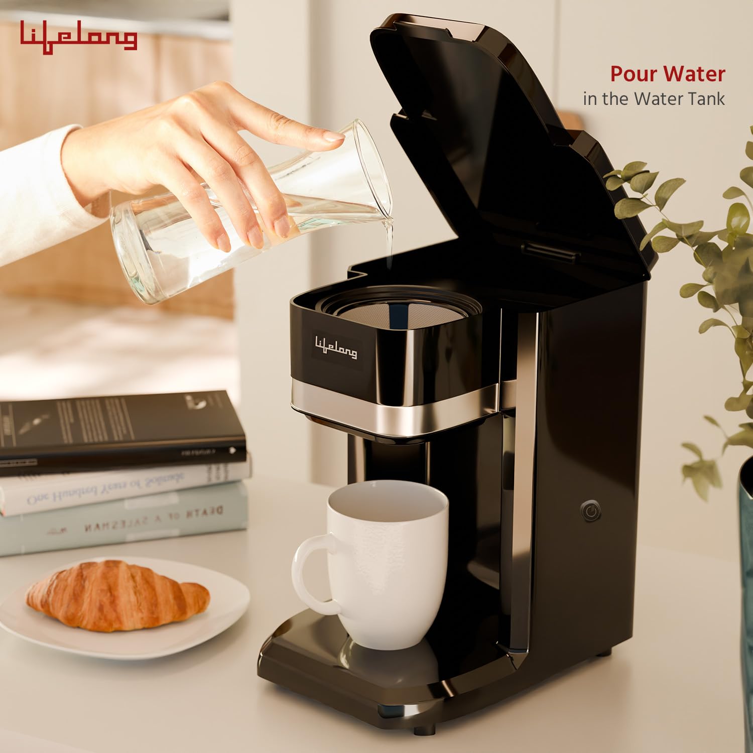 A black single-serve coffee maker with a white mug underneath, shown in a kitchen-like setting with a hand pouring water from a glass into the tank. A croissant and books sit on a plate beside it, and the brand name 'Lijelong' is visible on the machine.