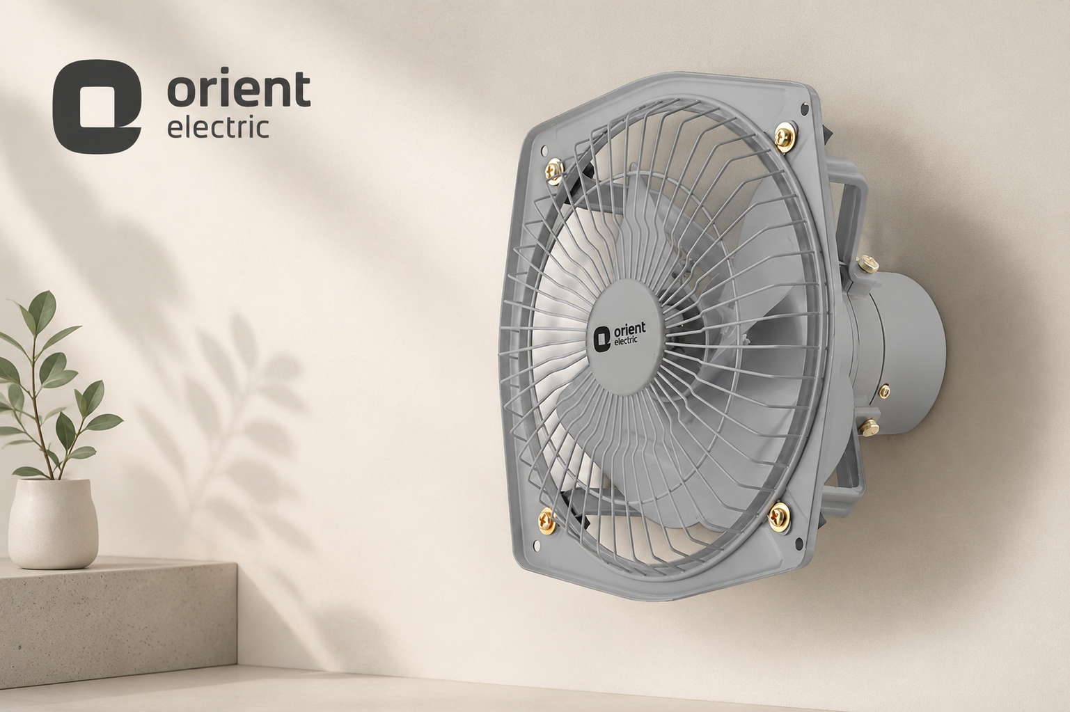 A wall-mounted gray electric fan with a protective grille, shown mounted on a beige wall. The Orient Electric brand logo is visible both on the fan and as an overlay in the top left corner.