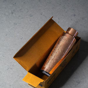 A copper-colored cylindrical bottle or container resting inside an open mustard-yellow cardboard box on a gray concrete surface.
