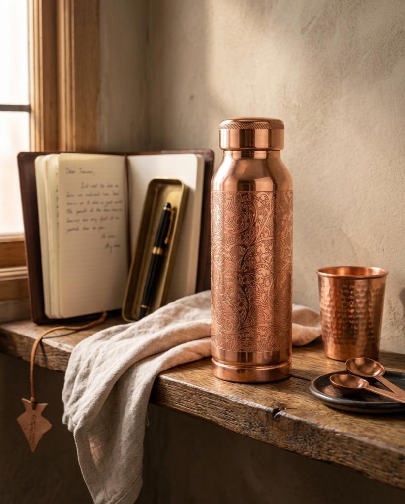 A copper-toned insulated bottle with ornate embossed floral patterns stands on a rustic wooden windowsill beside a matching hammered copper cup, spoon, and a folded cream cloth. An open notebook and pen case are visible in the background near a window.