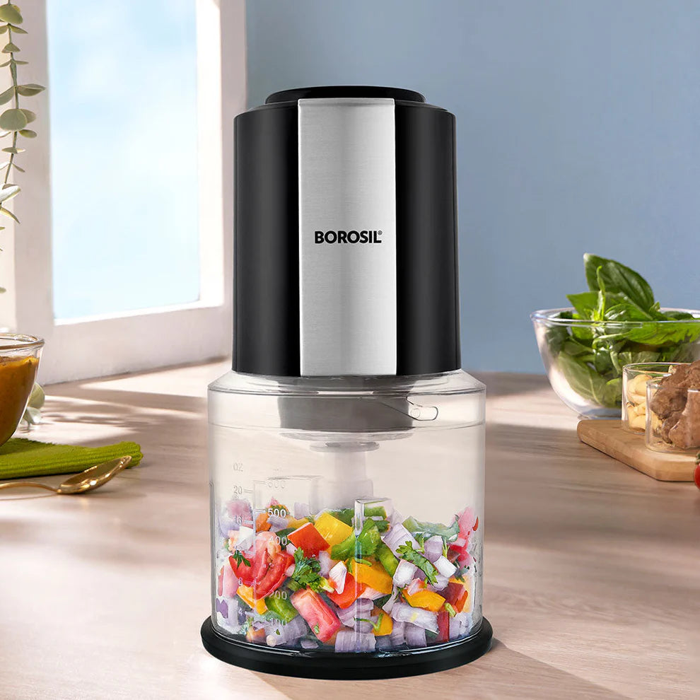 Borosil electric food chopper with a stainless steel motor housing and transparent bowl, finely chopping fresh onions, tomatoes, capsicum, and herbs. Compact and powerful kitchen appliance for quick vegetable preparation, meal prep, and everyday cooking.